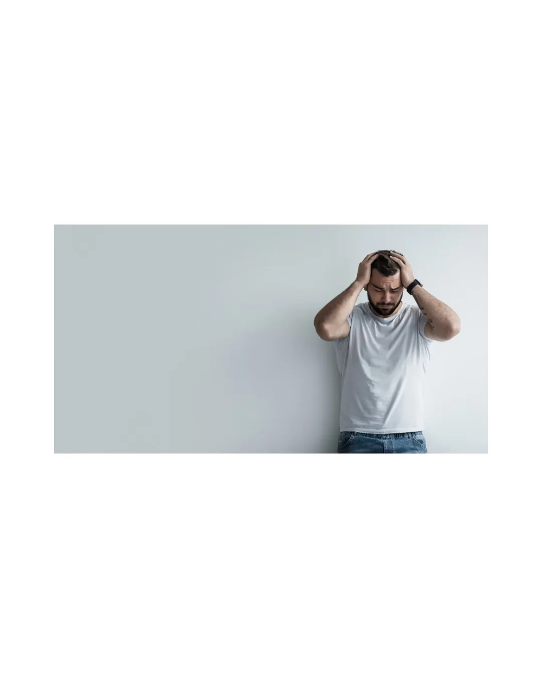 A man standing against a grey wall, hands on his head, looking distressed